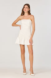A woman is wearing a strapless, white, fitted mini dress with horizontal ruching detail throughout the bodice and skirt. The dress features a ruffled hem at the bottom, adding a playful touch. She is also wearing white high-heeled shoes with ankle straps and embellishments.